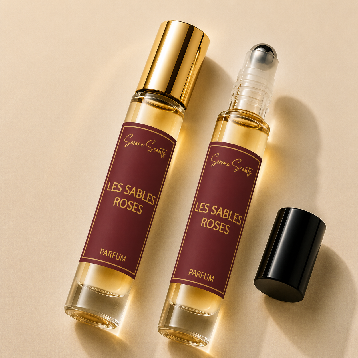 Two perfume roller bottles labeled 'Grève Scénary Les Sables Roses Parfum' with gold caps, shown on a warm beige background with one bottle uncapped and its black cap beside it.